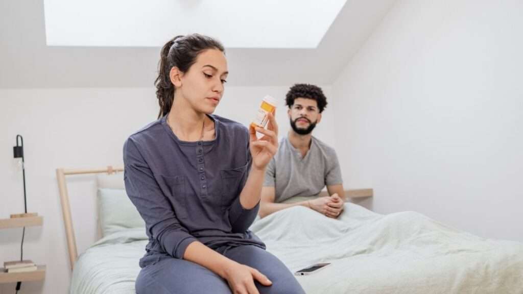 Woman sat in bed in front of a man holding the contraception pill